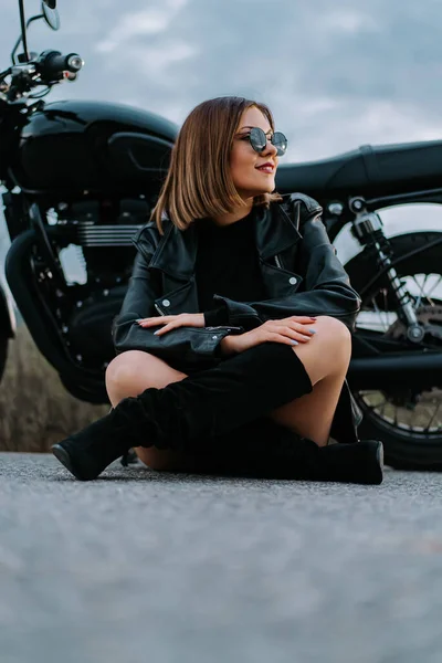 Motorcyklist kvinna i minikjol sitter på asfalten nära hennes retro-stylade motorcykel. Sexig kvinnlig förare i jackboots och solglasögon på vägen. Resa, hastighet, frihet koncept. — Stockfoto