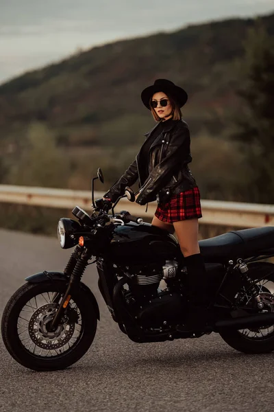 미니스커트를 입은 세련 된 모터사이클 선수 여성, 빈티지 스타일의 오토바이 위에 가죽 재킷을 입은 여성. 고속 도로에서 잭 부츠를 신은 매혹적 인 운전자. 트립, 속도, 스타일, 페미니즘 개념. — 스톡 사진