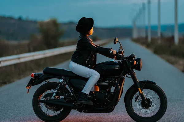 Hipster stylish motorcyclist woman sitting on vintage-styled motorcycle. Young female driver in hat at evening on roadway. Trip, freedom, classic motorbike concept. — Stock Photo, Image