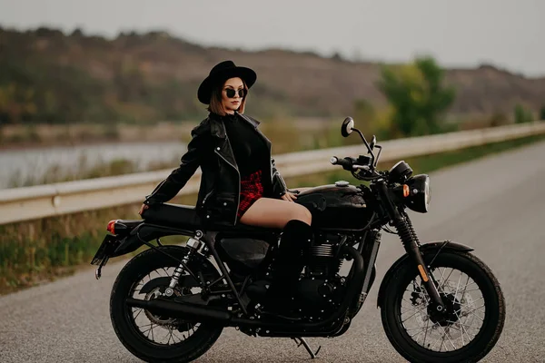 Mujer motociclista en mini falda sentada en motocicleta de estilo retro. Conductora sexy con botas de chaqueta en la carretera. Viaje, velocidad, concepto de libertad. —  Fotos de Stock