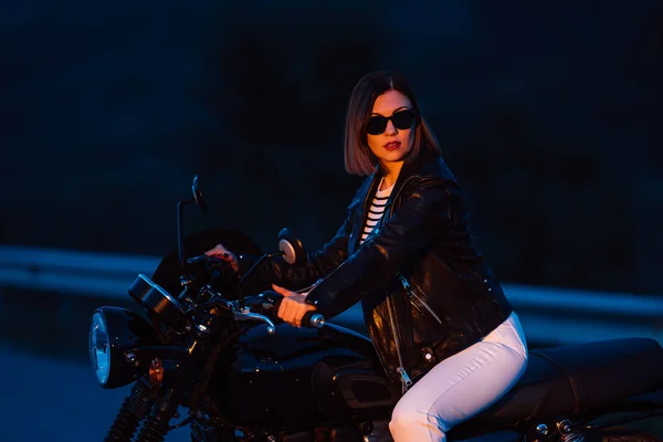 Hipster stilvolle Motorradfahrerin sitzt auf einem Motorrad im Vintage-Stil. Junge Autofahrerin nachts auf der Fahrbahn. Reise, Freiheit, klassisches Motorradkonzept. — Stockfoto