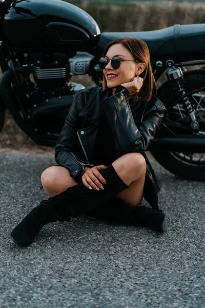 Mulher motociclista em mini-saia sentado no asfalto perto de sua motocicleta retro-styled. Motorista feminina sexy em botas e óculos de sol na estrada. Viagem, velocidade, conceito de liberdade. — Fotografia de Stock