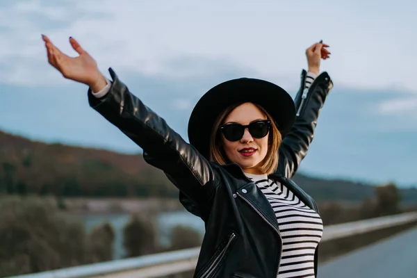 Ritratto di giovane donna spensierata in piedi sulla strada notturna vuota a braccia aperte. Ragazza in top a righe e giacca di pelle. Hipster col cappello. Elegante signora facendo gesto di libertà — Foto Stock