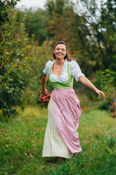 Щаслива жінка в довгій ретро-сукні з фартухом біжить з кошиком в яблучному саду. Органічний сільський спосіб життя, сільське господарство, урожай, концепція вінтажного стилю . — стокове фото