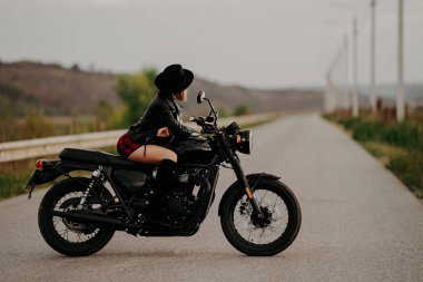 Motosikletli kadın mini etekli retro-stil motosikletin üzerinde oturuyor. Yoldaki çizmeli ve şapkalı seksi yabancı kadın sürücü. Trip, hız, özgürlük kavramı.