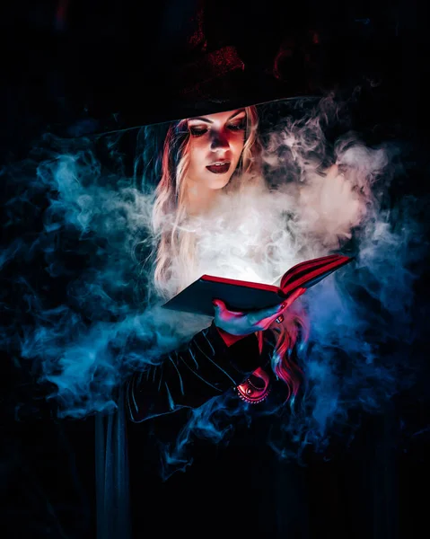 Belle sorcière lisant un livre magique sur fond sombre en forêt. Halloween, concept de costume de sorcier blonde. Jolie femme en fumée bleue. — Photo