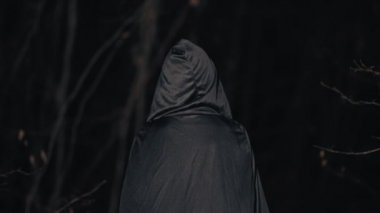 Karanlık bir gecede ormanda ağaçların arasında yürüyen siyah cadı olarak tanınmayan bir kadın. Uzun elbiseli, pelerinli bir kız. Cadılar Bayramı konsepti, ürkütücü kostüm