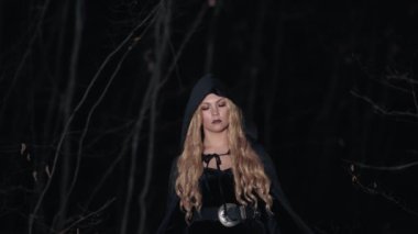 Kapüşonlu uzun sarı saçlı siyah cadı korkunç gece ormanlarında yürüyor. Uzun elbiseli ve paltolu kadın. Cadılar Bayramı, korku, konsept, ürkütücü kostüm