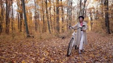 Sarı sonbahar parkında bisikleti olan klasik trençkotlu genç bir kadın. Doğanın arka planında trend bir kızın inanılmaz sahnesi. Çevre dostu ulaşım şekli