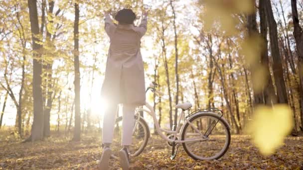 Ung kvinna i klassisk trenchcoat jublar höst, frihet och vacker natur. Flicka utspridda blad. Kärlek, tacksamhet koncept. Fantastisk scen av trendiga flicka med cykel. — Stockvideo