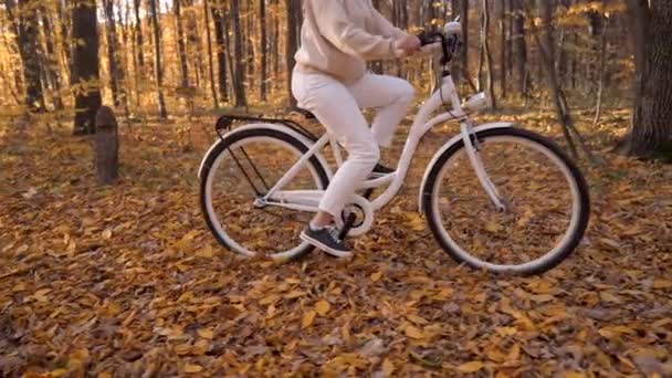 Bliższe nogi kobiety w białych spodniach jeżdżącej samotnie w jesiennym parku. Słoneczny dzień, złote liście w jesiennym żółtym lesie. Nierozpoznawalna dziewczyna na rowerze vintage, zdrowy styl życia, materiał estetyczny. — Wideo stockowe