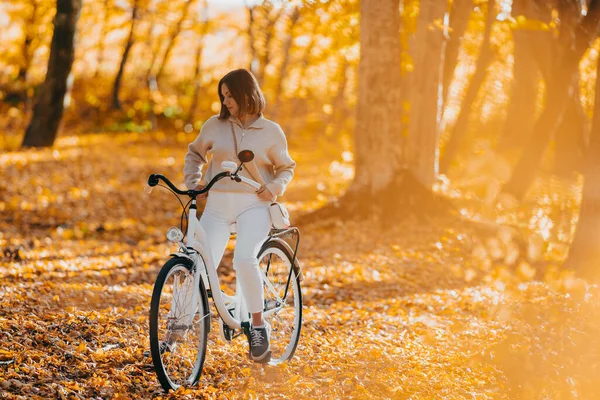 秋天公园里骑自行车的年轻女子的美丽画像。阳光灿烂的日子,金黄的树叶在森林里.穿着老式白色自行车的时髦女士，健康的生活方式，美观的场面 — 图库照片