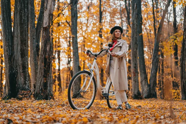 黄秋公园里穿着经典风衣、骑复古自行车的年轻女子。在大自然的背景下,迷人的时尚女孩的场景.无害环境的运输方式 — 图库照片