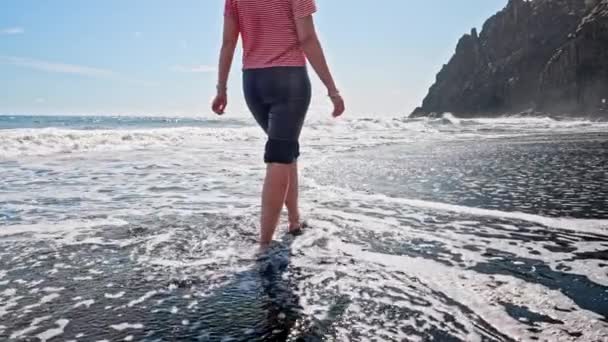 Πόδια γυναίκας περπατάνε ξυπόλητοι σε υγρή μαύρη ηφαιστειακή παραλία νησί άμμο. Όμορφα πόδια της νεαρής κοπέλας κοντά στη θάλασσα κατά το ηλιοβασίλεμα ή την ανατολή. Αργή κίνηση. — Αρχείο Βίντεο