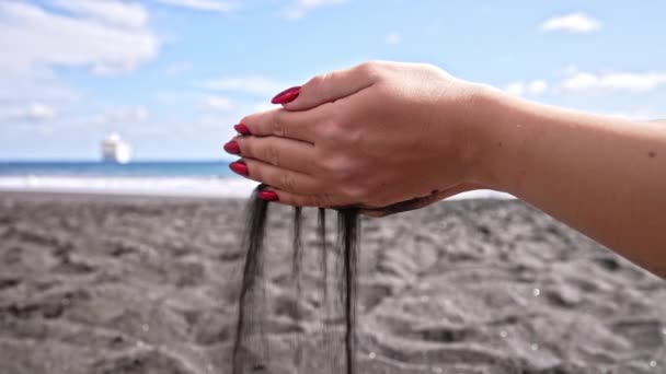 Kobieca ręka wylewająca wulkaniczny czarny piasek płynący przez palce w zwolnionym tempie na wyspie tropikalnej plaży. Piękna scena młodej kobiety z czerwonymi paznokciami bawiącej się ziarnem na zachodzie słońca. — Wideo stockowe