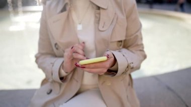 Şehir caddesinde akıllı telefon kullanan güzel bir kadın. Merkez meydandaki çeşmede tek başına oturan bir kadın, mobil cihazla internette sörf yapıyor. Sonbahar sezonu. Teknoloji, sosyal uygulamalar, bağlantı kavramı