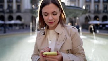 Şehir caddesinde akıllı telefon kullanan güzel bir kadın. Merkez meydandaki çeşmede tek başına oturan bir kadın, mobil cihazla internette sörf yapıyor. Sonbahar sezonu. Teknoloji, sosyal uygulamalar, bağlantı kavramı