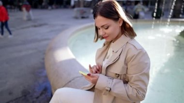 Şehir caddesinde akıllı telefon kullanan güzel bir kadın. Merkez meydandaki çeşmede tek başına oturan bir kadın, mobil cihazla internette sörf yapıyor. Sonbahar sezonu. Teknoloji, sosyal uygulamalar, bağlantı kavramı