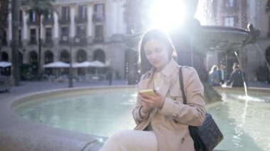 Şehir caddesinde akıllı telefon kullanan güzel bir kadın. Merkez meydandaki çeşmede tek başına oturan bir kadın, mobil cihazla internette sörf yapıyor. Sonbahar sezonu. Teknoloji, sosyal uygulamalar, bağlantı kavramı