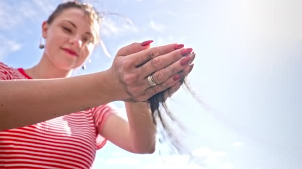 Femme souriante versant du sable noir volcanique traversant les doigts au ralenti sur la plage tropicale de l'île. Belle scène de jeune femme avec des ongles rouges jouant avec du grain au coucher du soleil. — Video