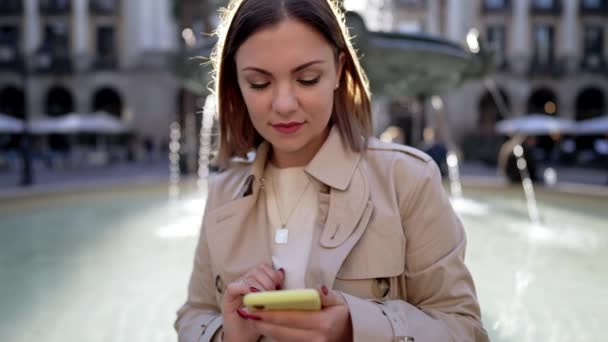 도시의 거리에서 스마트폰을 사용하는 예쁜 여성. 중앙 광장의 분수에 홀로 앉아 있는 여성은 모바일 기기로 인터넷 서핑을 합니다. 가을 시즌이다. 기술, 소셜 앱, 연결 개념 — 비디오
