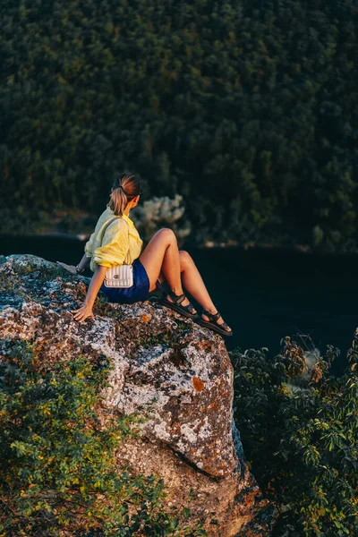 Young stylish woman sits on rock near a cliff. Hiking trip. Amazing nature, summer and sunset. Unrecognizable travel girl. Adventure lifestyle — Stock Fotó