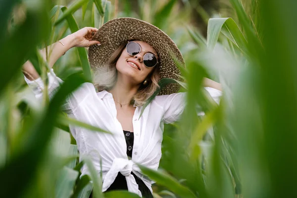 Portret młodej stylowej kobiety w zielonej dżungli. Dziewczyna w słomkowym kapeluszu i okularach przeciwsłonecznych, pościel. Pani wygląda na szczęśliwą i zdrową, uśmiecha się. — Zdjęcie stockowe