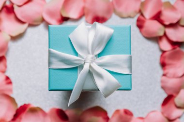 Gül yapraklarıyla çerçevelenmiş beyaz arka planda ipek yay ile bağlanmış romantik hediye mavi kutu. Yıldönümü hediyesi, kartlar, sevgililer günü kartpostalları, duvar kağıdı konsepti..