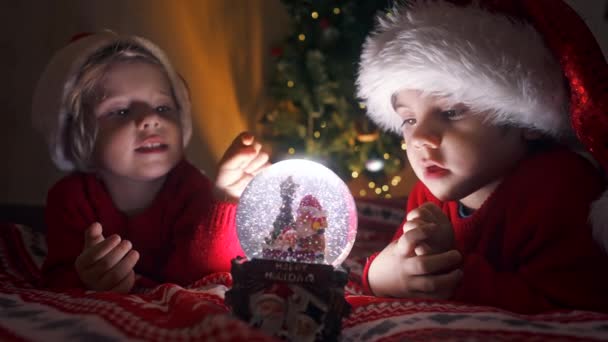Lindos niños en sombreros de Santa con bola de nieve. Juguete de regalo de Año Nuevo para niños. Tiempo mágico de Navidad, los niños pequeños miran la bola de vidrio con interés. Familia, hermanos, vacaciones y concepto de celebración — Vídeos de Stock