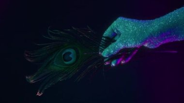 Kadın eli, neon renkli ışığın altında tavus kuşu tüyü tutan holografik parıltılarla kaplı. Vücut sanatı, görkemli kavramsal çekim, moda tarzı.. 