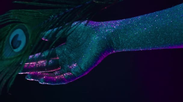 Peacock φτερό χαϊδεύει θηλυκό χέρι που καλύπτεται με ολογραφικό λαμπερό glitter κάτω από νέον χρωματιστό φως. Ζωγραφική σώματος, λαμπερό εννοιολογικό υλικό, — Αρχείο Βίντεο