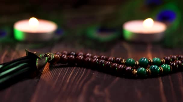 Dolly shot of beautiful wooden mala beads with green gemstones on table decorated with candles and peacock feathers. Meditation, mantra, spirituality world, religion and God concept. — Stock Video