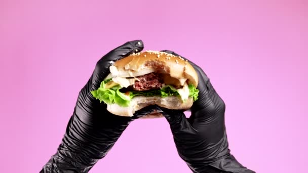 Mãos em luvas de látex preto com hambúrguer mordido no fundo rosa. Mulher comendo patty apetitoso com costeleta de carne, legumes, queijo derretido e maionese. Conceito de fast food. — Vídeo de Stock