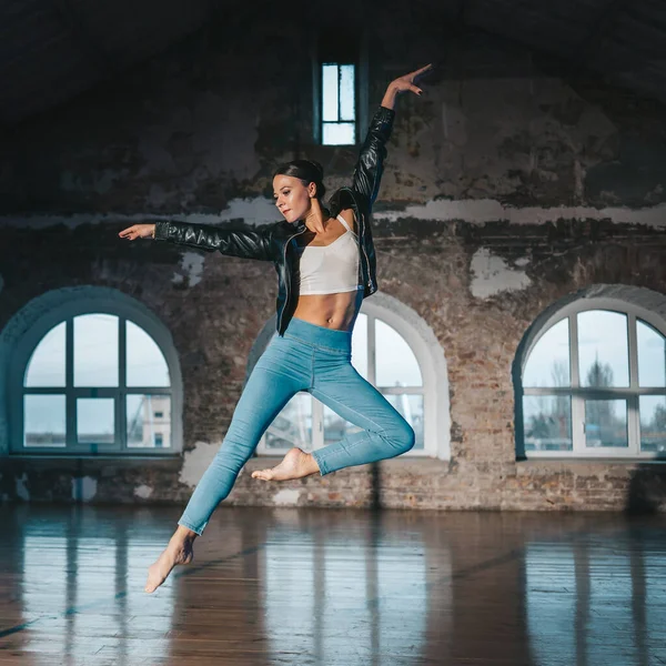 Wanita muda melompat dengan gaya kasual jeans dan jaket kulit melakukan balet di studio tua. Praktik balerina yang menarik dalam koreografi saja — Stok Foto