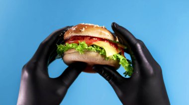 Elleri siyah eldivenlerle mavi arka planda burger tutuyor.