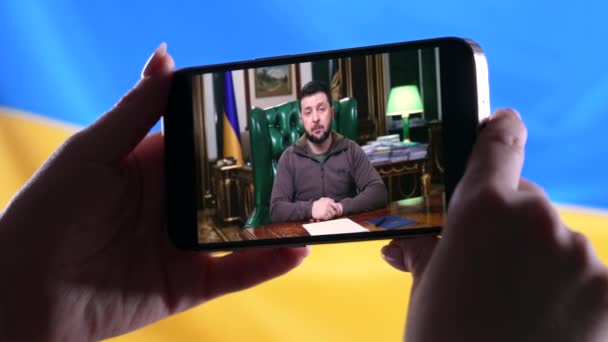 Απρίλιος 2022 - Κίεβο, Ουκρανία. Παρακολουθώντας online ομιλία του Προέδρου Volodymyr Zelensky σε smartphone με φόντο την ουκρανική σημαία. Ειδήσεις. Ρωσική επίθεση. Απειλή πολέμου. — Αρχείο Βίντεο