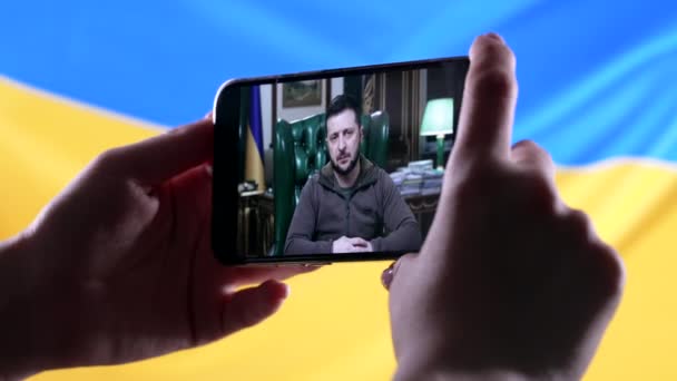 Aprile 2022 Mariupol, Ucraina. Guardando il discorso online del presidente Volodymyr Zelensky su smartphone su sfondo bandiera ucraina. Notizie. Aggressione russa. Minaccia di guerra. — Video Stock