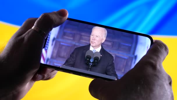 Abril de 2022 - Kharkiv, Ucrania. Ver el discurso en línea del presidente Joe Biden en el teléfono inteligente sobre fondo de bandera ucraniana. Noticias. Agresión rusa. Amenaza de guerra. — Vídeo de stock