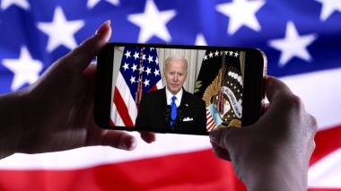Nisan 2022 - New York, ABD. Başkan Joe Biden 'ın çevrimiçi konuşmasını Amerikan bayrağı arka planında akıllı telefondan izliyorum. Ukrayna ve Rusya arasındaki gerginlikler hakkında haberler.