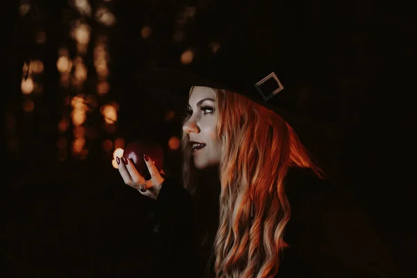 Sorcière blonde en noir tenant une pomme rouge la nuit en forêt. Halloween, concept de sorcier effrayant. Superbe portrait de femme. symbole de tentation, poison. Conte de fées neige blanche concept — Photo