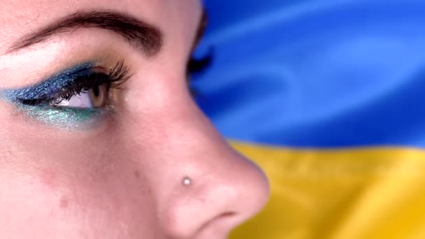 Жіноче обличчя, очі з патріотичним українським синьо-жовтим макіяжем. Блискучі тіні і довгі вії. Жінка зелене око на фоні прапора — стокове відео