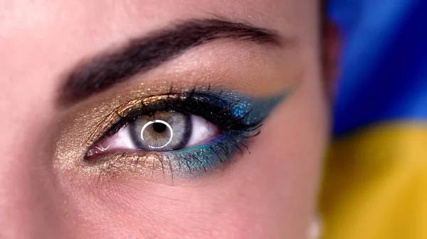 Macro view of woman eye with blue yellow shiny make-up. Lady, green cornea with round reflection. Ukrainian model on flag background. — ストック写真