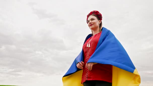 Smiling ukrainian woman with national flag on sky background. Portrait iof young lady in red embroidery Vyshyvanka and flowers wreath. Ukraine freedom, patriot symbol, victory in war. — Wideo stockowe