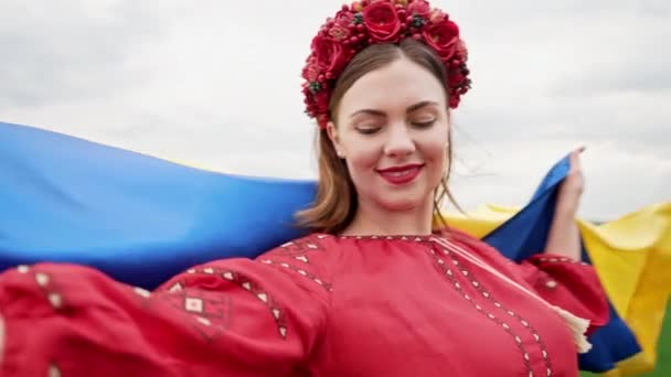 Happy ukrainian woman spinning around with national flag on sky background. Lady in red embroidery Vyshyvanka and flowers wreath. Ukraine freedom, patriot symbol, victory in war. — Wideo stockowe