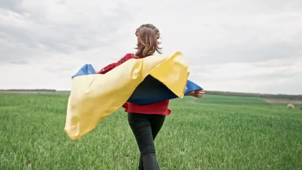 Feliz mujer ucraniana corriendo con bandera nacional al aire libre en el campo verde. Señora en bordado rojo Vyshyvanka y flores corona. Ucrania libertad, símbolo patriota, victoria en la guerra. — Vídeo de stock