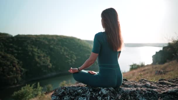 Unrecognizable concentrated woman in lotus pose meditating on high rock above water. Calm yoga concept, zen, relaxation, practice on nature background. — ストック動画