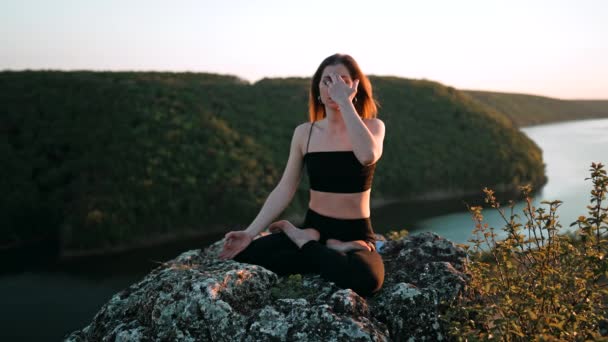 Young woman doing yoga belly exercise - Kapalabhati yogic breathing technique. Yogi detoxtechnique while sitting in lotus pose on high cliff above water outdoors. Lady slows breathing and meditating. — Video Stock