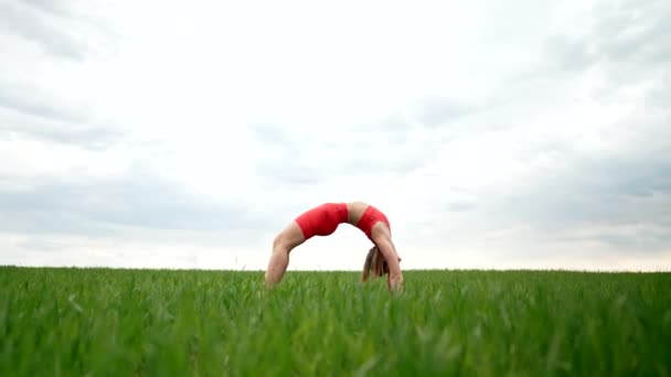 Young woman in orange practicing yoga in nature, green field. Girl makes bridge. Concept of fitness, sports, healthy lifestyle. — Stok video