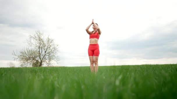 Sporty woman in orange wear doing yoga in fresh green field. Complex of asanas - Surya Namaskar, balance, zen. Fitness, everyday practice on nature, healthy lifestyle concept. — Wideo stockowe