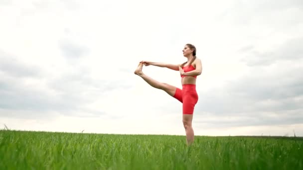 Woman in orange sports wear practicing yoga - utthita hasta padangushthasana in green field. Balance on one leg. fitness, sport, healthy lifestyle concept. — Video Stock